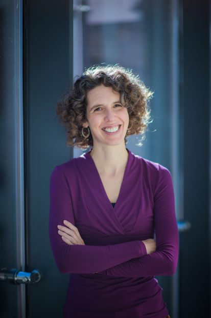 Headshot of white female wearing purple blouse standing against a window with steel gray columns