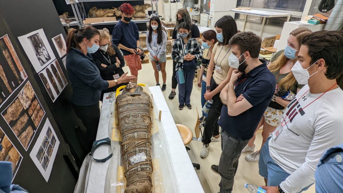 A group of students standing over a table with an ancient tomb listening to a female presenter discuss the ancient artifact