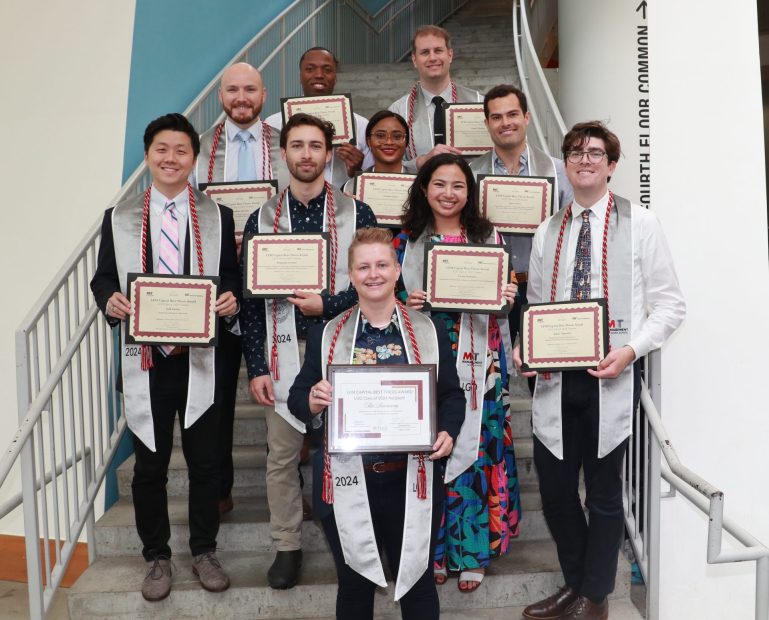 A group of graduates pose on stairs holding awards.