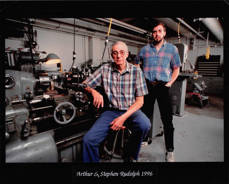 One older man sitting, with his son standing behind him in a machine shop.